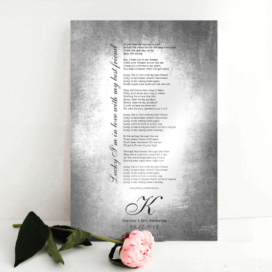 Couples custom song lyrics wall decor - vintage metal tin aluminum song plaque sits on a white background with a single pink rose. The sign features the inscription “lucky I’m in love with my best friend” in an elegant calligraphy running vertically up the side of song lyrics in a stanza format - which is an except from the lyrics. Beneath is a last name Monogram letter, names and a union date, for posterity. Photo courtesy of Honeycomb Proverbs