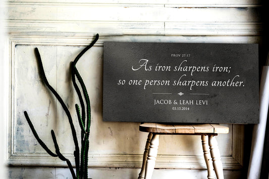 Wall art shown, set on the seat of a rustic wooden chair. It's a dark grey, distressed metal sign featuring the popular iron-themed bible verse, Prov. 27:17, "As iron sharpens iron so one person sharpens another." The words of the verse take center stage, elegantly scripted in a modern calligraphy font, and a delicate accent beneath. A personalization or dedication sits below the accent: the couple's first names and union date.