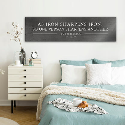Metal sign with silver text that reads "As iron sharpens iron; so one person sharpens another." The sign also includes the names "Bob & Jessica" and the scripture reference "Proverbs 27:17".