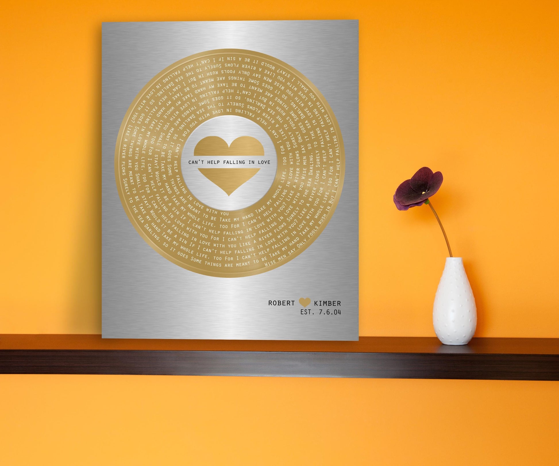Heart accented, metallic mixed metal tin aluminum and brass sign with custom song lyrics, silver metal with brass vinyl record motif with inscribed song, and personalized with names and a date. The vinyl record sits prominently and is centered for a contemporary mix of silver and gold metals. The personalized metal plaque is sitting on a ledge next to a vase.