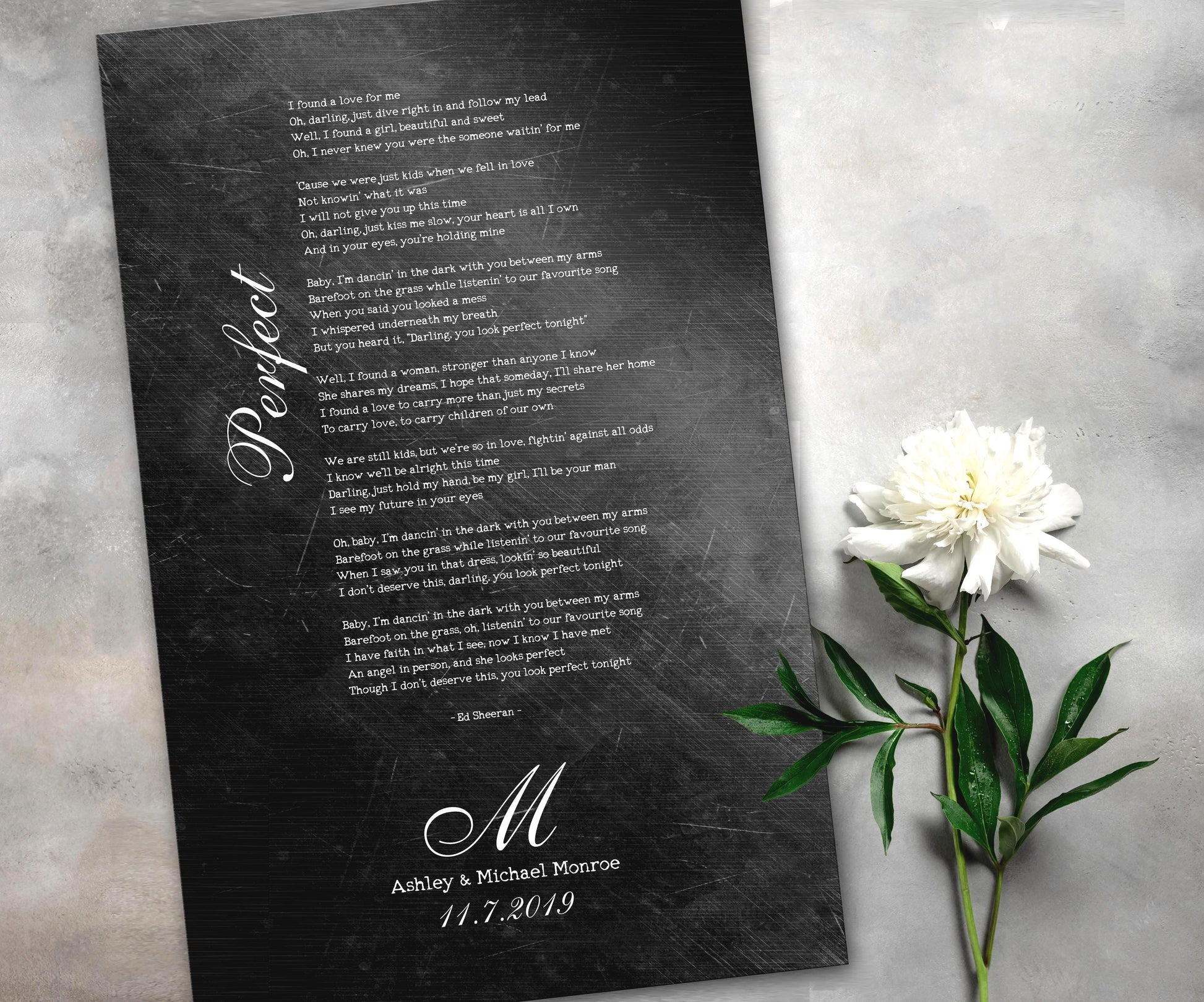 Iron anniversary gifts like this distressed metal song plaque featuring personalized song lyrics, a couple's name and anniversary date are becoming very popular for husband and wife anniversaries. The rustic and distressed affect give this keepsake a rugged feel while also blending a touch of elegance and whimsy with it's thoughtful font pairing of vintage type writer and calligraphy. A white rose sits beside the plaque which is laying on a table top.