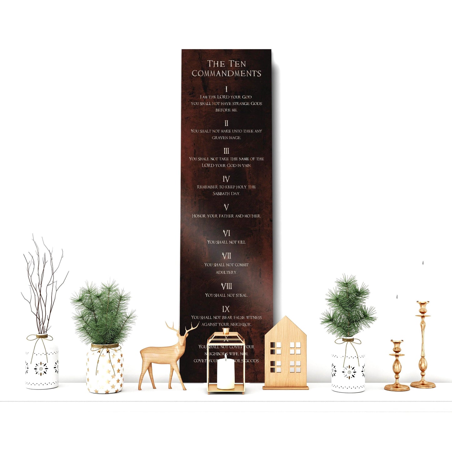 May include: A rustic rubbed bronze metal sign with the Ten Commandments written in white text. The sign is hanging on a white wall with a decorative shelf below it. The shelf has a white vase with a green plant, a white vase with a brown plant, a gold candle holder with a white candle, a gold candle holder with a white candle, a wooden house decoration, and a gold deer figurine.