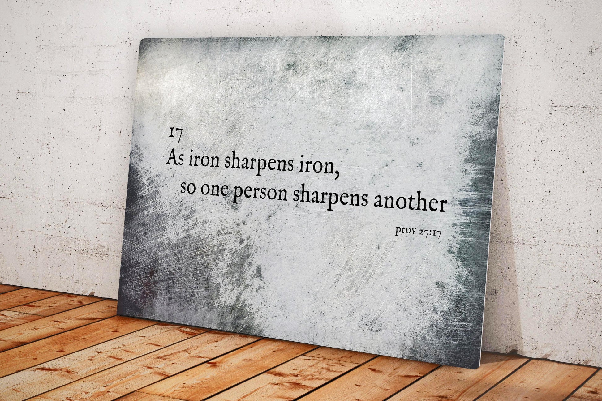 Metallurgy Christian wall decor: A tarnished metal sign with the verse, Proverbs 27:17, "As iron sharpens iron, so one person sharpens another".
Sign sits on wood plant floor and is leaning against a concrete wall.
Courtesy of Honeycomb Proverbs