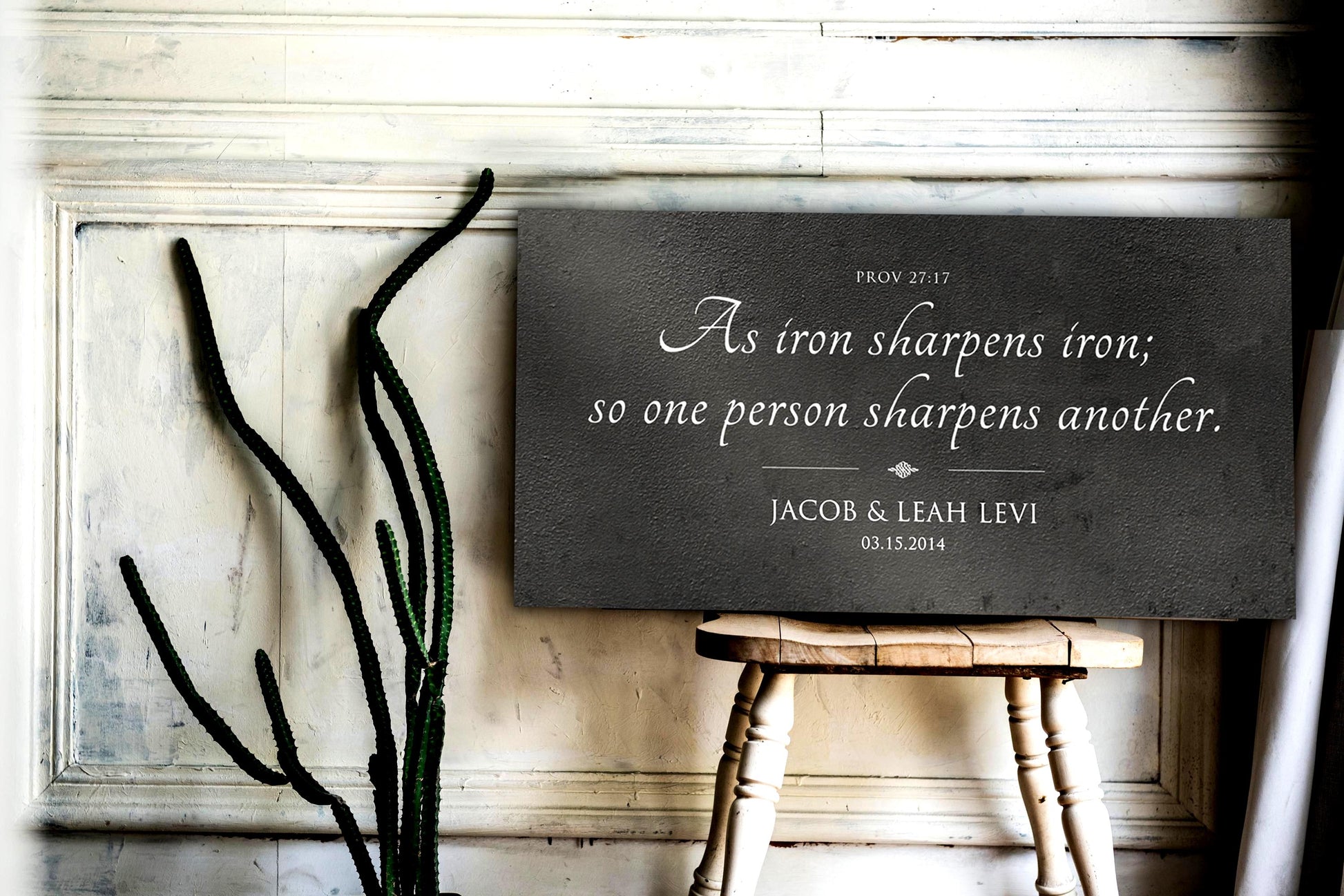 Wall art shown, set on the seat of a rustic wooden chair. It's a dark grey, distressed metal sign featuring the popular iron-themed bible verse, Prov. 27:17, "As iron sharpens iron so one person sharpens another." The words of the verse take center stage, elegantly scripted in a modern calligraphy font, and a delicate accent beneath. A personalization or dedication sits below the accent: the couple's first names and union date.