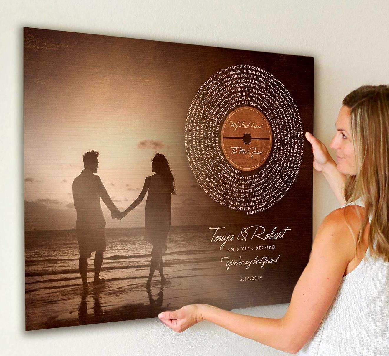 An attractive woman is hanging a bronze anniversary plaque on her wall. This anniversary Photo Gift and burnished bronze keepsake blends a favorite photo with a vinyl record-inspired motif, displaying the full song lyrics in a striking spiral. Below the record, an excerpt from the lyrics, years married, couples names, and est. date complete this timeless piece.
