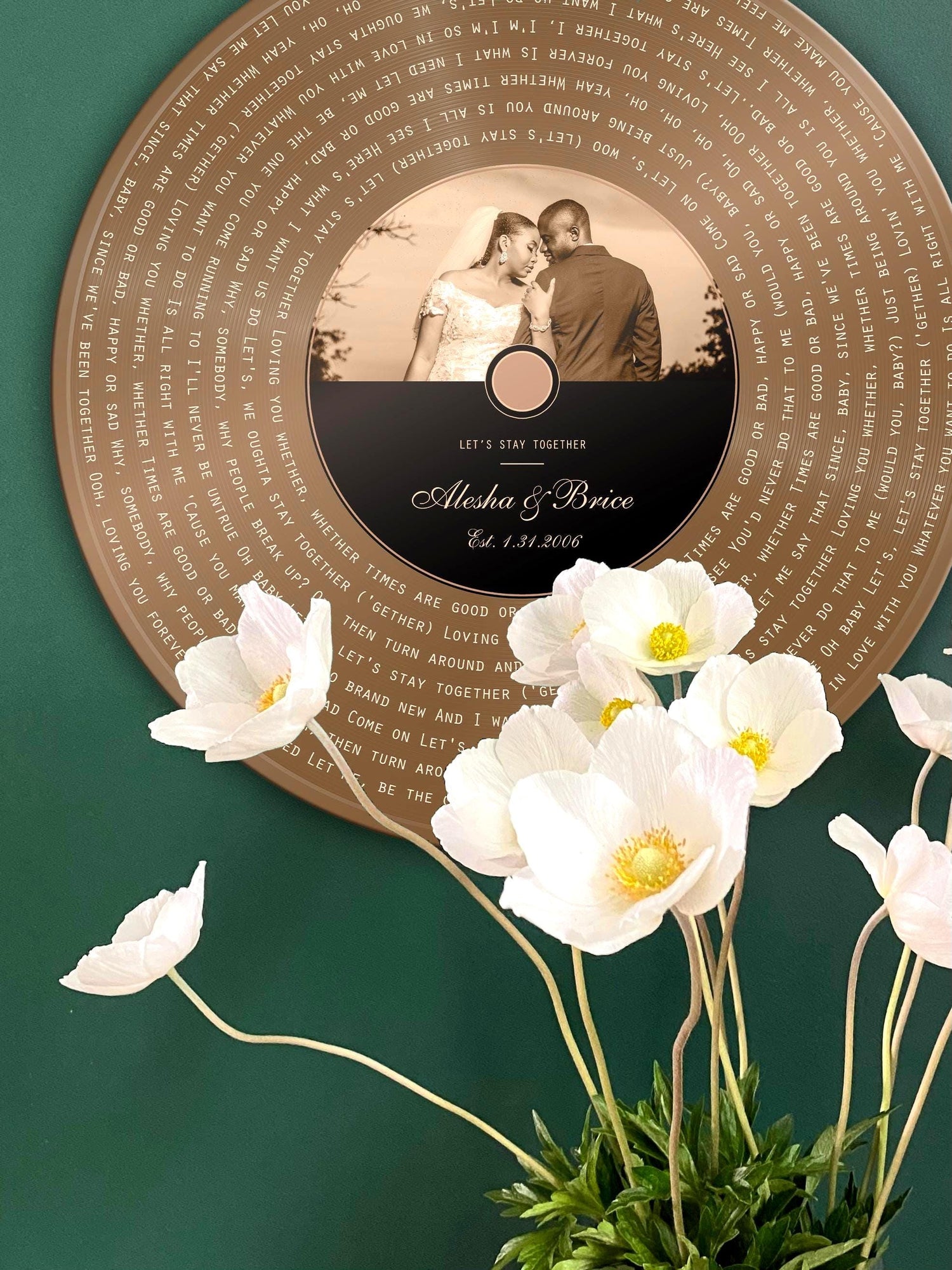 Personalized Bronze Anniversary record includes song lyrics of choice, overlaid onto the vinyl motif and photo and song credit with the couple's names are inscribed onto the record label area.
Round Bronze metal record is hung on a green wall and white flowers delicately frame the lower third of the photo.

Photo courtesy of Honeycomb Proverbs
