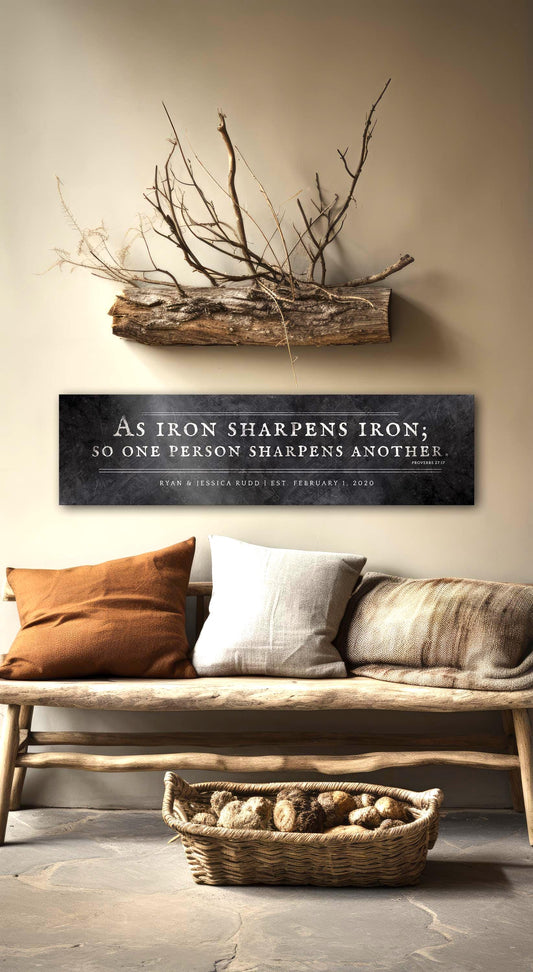 Distressed Iron Alloy As Iron Sharpens Iron Sign This wide format wall decor features the words of Proverbs 27:17inscribed in silver on a distressed dark wrought iron finish with an option to add a short personalization or dedication beneath the scripture. The sign is wall mounted above a sofa and is personalized with the names of a married couple and their union date. Photo courtesy of Honeycomb Proverbs