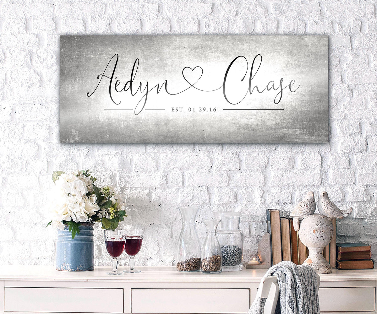 Unique Anniversary gifts like Honeycomb Proverbs' Personalized Heart Name signs are popular for ten year wedding anniversaries. One of their tarnished metal Heart Name sign hangs above an accent table. The sign is extra large sized and features personalized names connected by a heart in a whimsical calligraphy font. The couple's established date sits beneath their names to mark their special day.