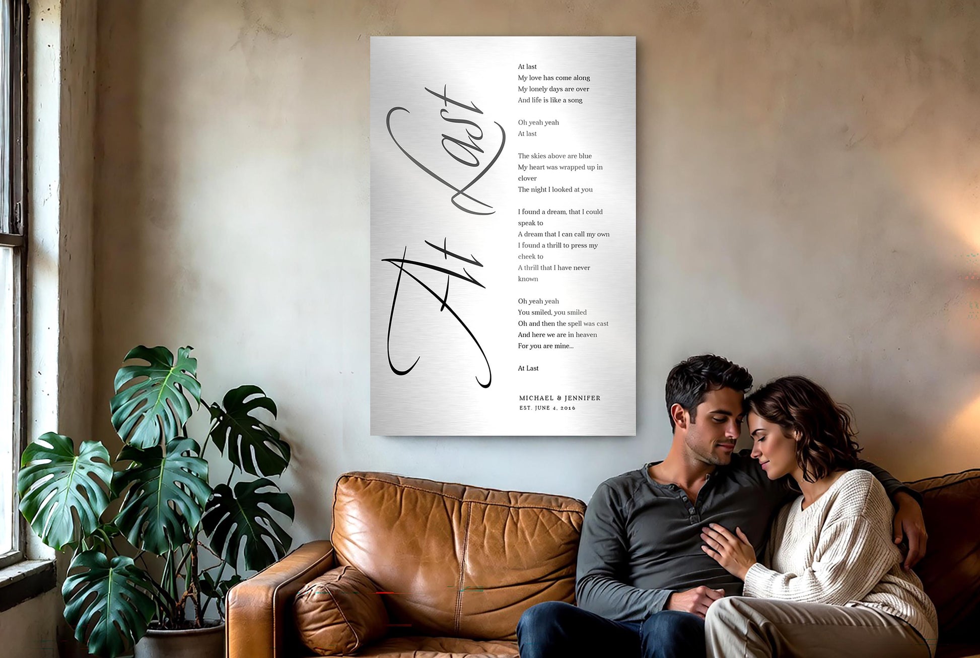 Large Custom Metal sign with wedding song lyrics "At Last" is displayed on wall. Seated slightly to the right of the frame, on a leather sofa is a couple in a loving embrace.