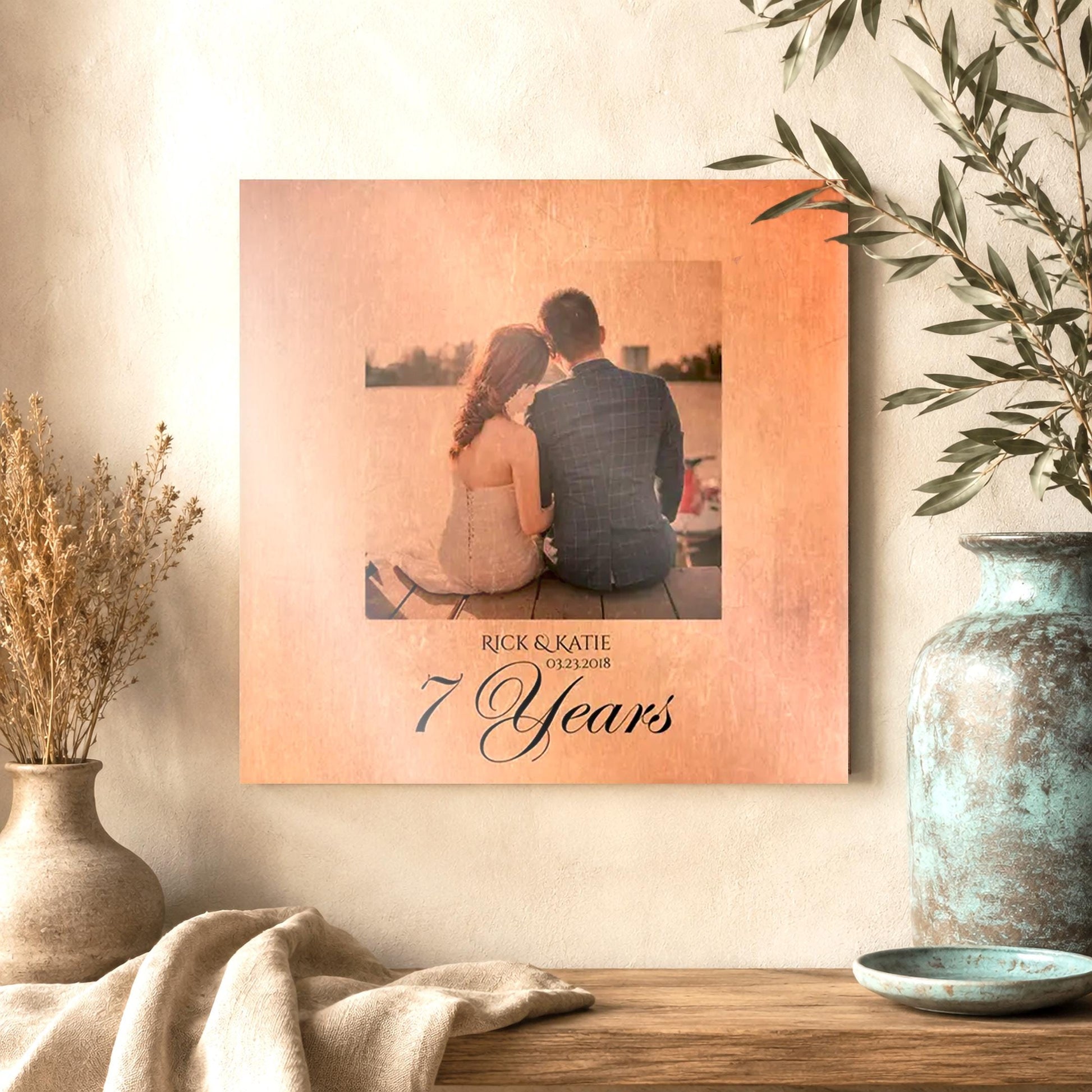 Beautiful Square Wall Decor on copper metal with "7 Year"s, and the names Nicole, and Robert, and the date 11.02.11"
A cropped portrait sits center, above the personalized inscription, and appears to also be in copper, with slight color tones showing through the copper base of the metal. The photo is of a bride and groom sitting together on a dock, shot from behind them with the water and mountains in the background. The artwork hangs on the wall of an elevated home space.