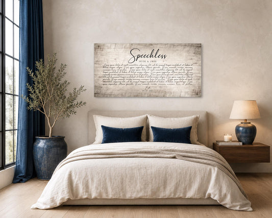 Modern neutral toned bedroom setting with a custom wooden song wall decor hanging above the bed, with the word, Speechless, in large calligraphy, and the names, Nick & Jess, beneath. There appear to be song lyrics taking up the main portion of the artwork beneath the names, and the abreviation "Est." sits beneath the paragraph of song lyrics, and a date reflecting the wedding date of Nick & Jess is inscribed at the very bottom of the wood wall art. The wood is an aged natural birch ton