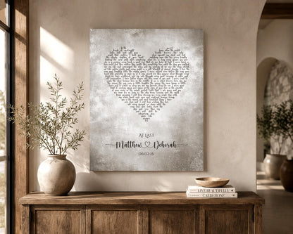 A tarnished metal artwork with custom lyrics shaped into a heart hanges on a wall near a window. The artwork is also personalized with the names of a married couple inscribed in a whimsical calligraphy font and joined by a decorative heart. Their union date sits beneath.  The song featured is At Last.