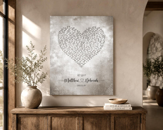 A tarnished metal artwork with custom lyrics shaped into a heart hanges on a wall near a window. The artwork is also personalized with the names of a married couple inscribed in a whimsical calligraphy font and joined by a decorative heart. Their union date sits beneath.  The song featured is At Last.