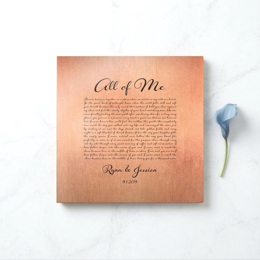 A copper metal song plaque sits on a marble surface with a blue lily accenting the right side of the frame. The song name is All of Me, and the plaque is finished with the names of a married couple and their union date, beneath a paragraph of song lyrics. The inscription is in black lettering in a calligraphic style letterhead.