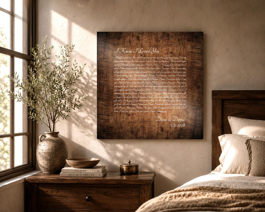 Wooden Song Lyrics Wall Art on a Stained birch square panel, with song lyrics inscribed with a calligraphy font. The song lyrics art is also personalized with the name of a married couple and their established date. It hangs on a wall in a rustic bedroom setting and sunlight comes through the window and landing on the wood artwork.