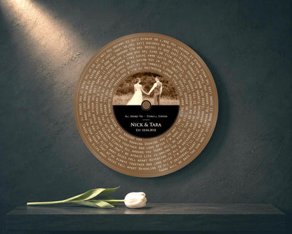 Bronze round song lyrics record with sepia couple photo and Nick and Tara label on a dark teal wall, white tulip on the shelf